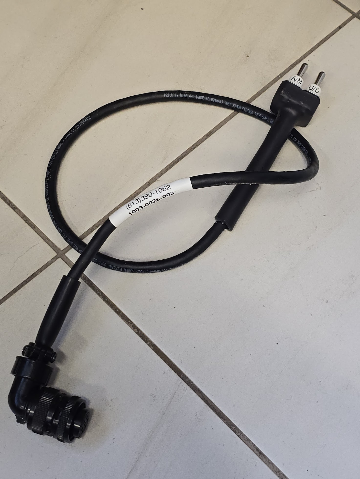 Black electrical cord with a right-angle connector and a plug, resting on a tiled surface. It may be used for powering devices or appliances.